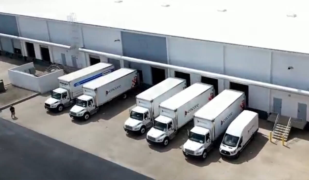 Aerial view of five white delivery trucks parked at loading docks outside a large industrial warehouse building, with a person walking nearby on the left.